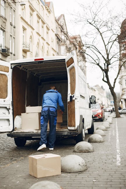 A man in a blue jacket and dark trousers is inside the back of a white commercial van parked on a city street, with the rear doors open during a home relocation process. The van is situated on cobblestone paving, with several large, rounded concrete bollards lining the roadside. The interior of the van contains wrapped furniture pieces, including a box-shaped item covered in white protective material. Outside the van, a cardboard box is placed on the pavement, ready to be loaded or unloaded. Additional parked cars and multi-storey residential buildings with classic architecture are visible in the background, along with leafless trees indicating a colder season. The scene captures the loading process for a furniture transport or moving service, likely coordinated by Man and a Van Dulwich, as part of packing, moving, and home relocation activities in Dulwich SE21.
