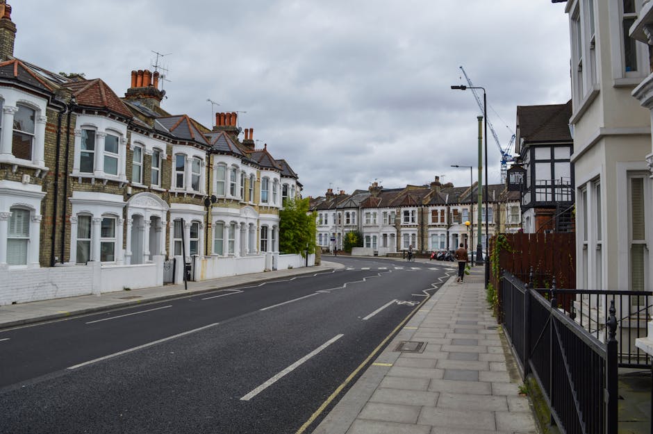 A quiet residential street scene in Dulwich featuring a row of Victorian-style terraced houses with white painted facades, bay windows, and red tiled roofs. The street has a paved sidewalk with black
