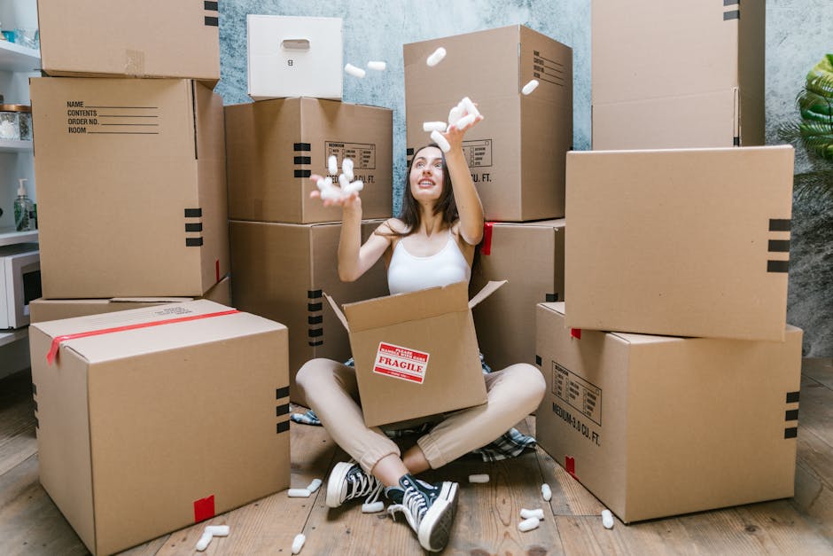 A young woman with dark hair sitting cross-legged on a wooden floor inside a room, surrounded by stacked cardboard boxes of various sizes for house relocation. She is wearing a white tank top, beige pants, and black-and-white sneakers, and is smiling while throwing white packing peanuts into the air. One open box with a red 'Fragile' sticker is positioned in her lap, and a few packing materials and tape are visible around her. The background includes a blue-textured wall, a small potted plant, and part of a kitchen or storage area, with natural lighting highlighting the scene. This image illustrates the packing and loading process involved in home removals, often handled by companies like Man and a Van Dulwich, who specialise in furniture transport and moving logistics.