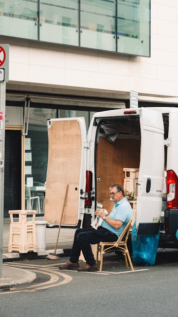A busy street scene in Dulwich featuring a variety of furniture, cardboard boxes, and packing materials arranged near the pavement and sidewalk, with some items being carried or prepared for loading. Visible are a few moving blankets, plastic wrap, and boxes stacked close to a white van used for home relocation purposes. Several individuals are seen walking along the street, engaged in activities related to moving or shopping. The background includes historic buildings and a clock tower with a weather vane on top, under a partly cloudy sky. The scene captures the logistics of furniture transport and packing during a same-day removal process, with the van parked nearby on the street in front of the shops, consistent with professional removal operations by Man and a Van Dulwich.